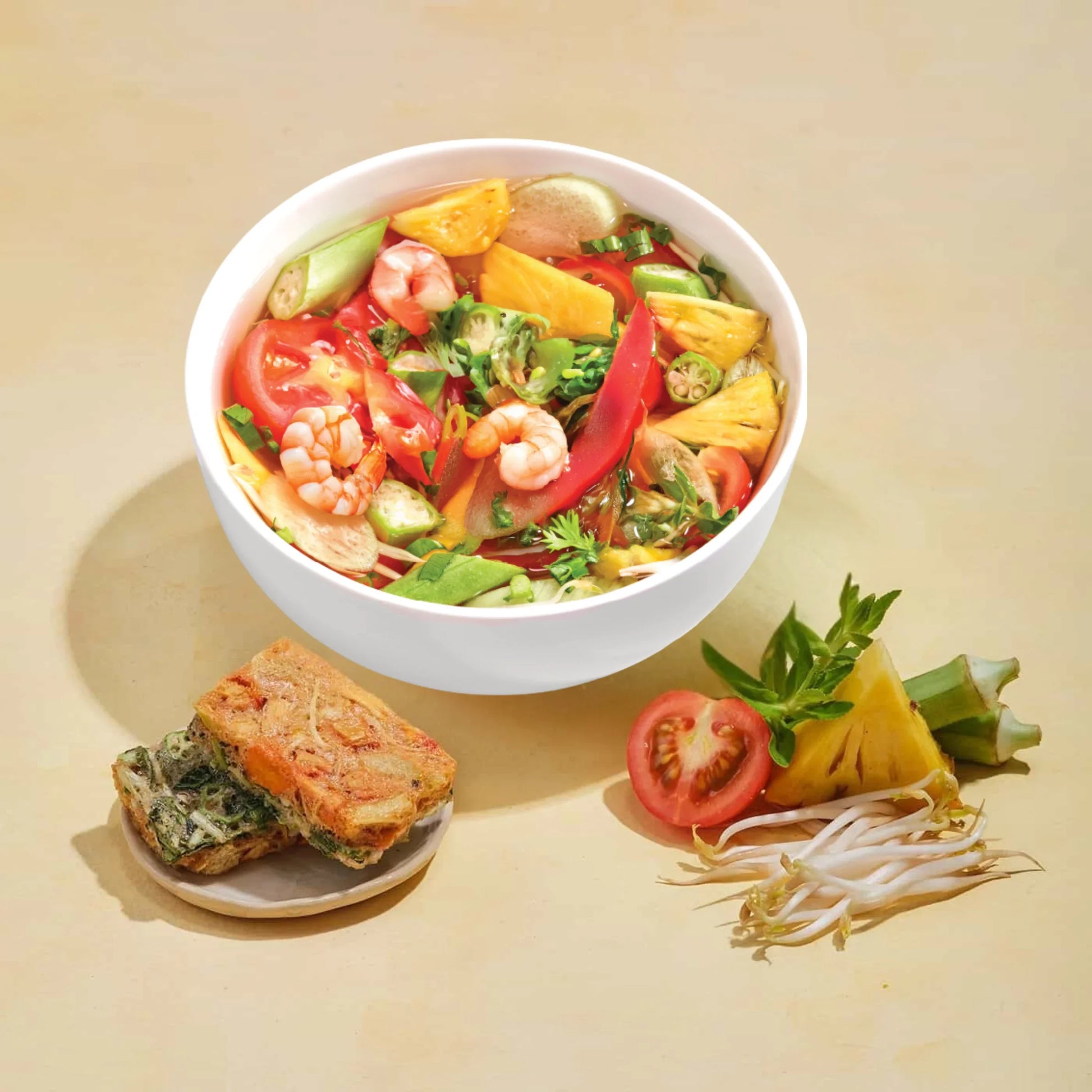 Bowl of shrimp soup with tomatoes, pineapple, okra, herbs, and side of vegetable fritters and fresh ingredients