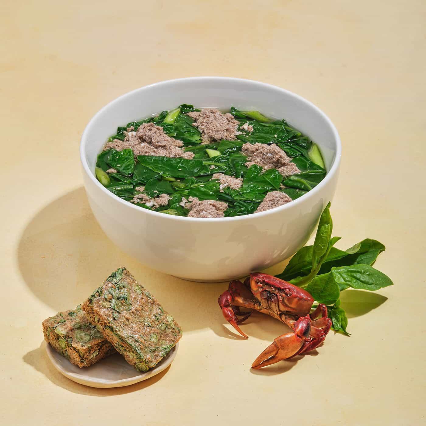 White bowl of spinach soup with ground meat, crab shell, fresh spinach, and spinach cakes on plate