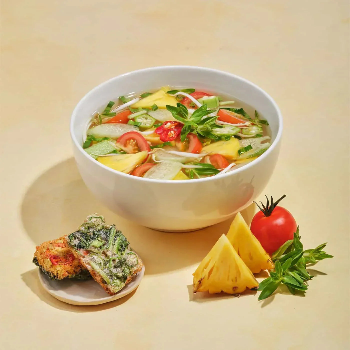 White bowl of vegan vegetable soup with okra, tomato, pineapple and basil, served with vegetable fritters